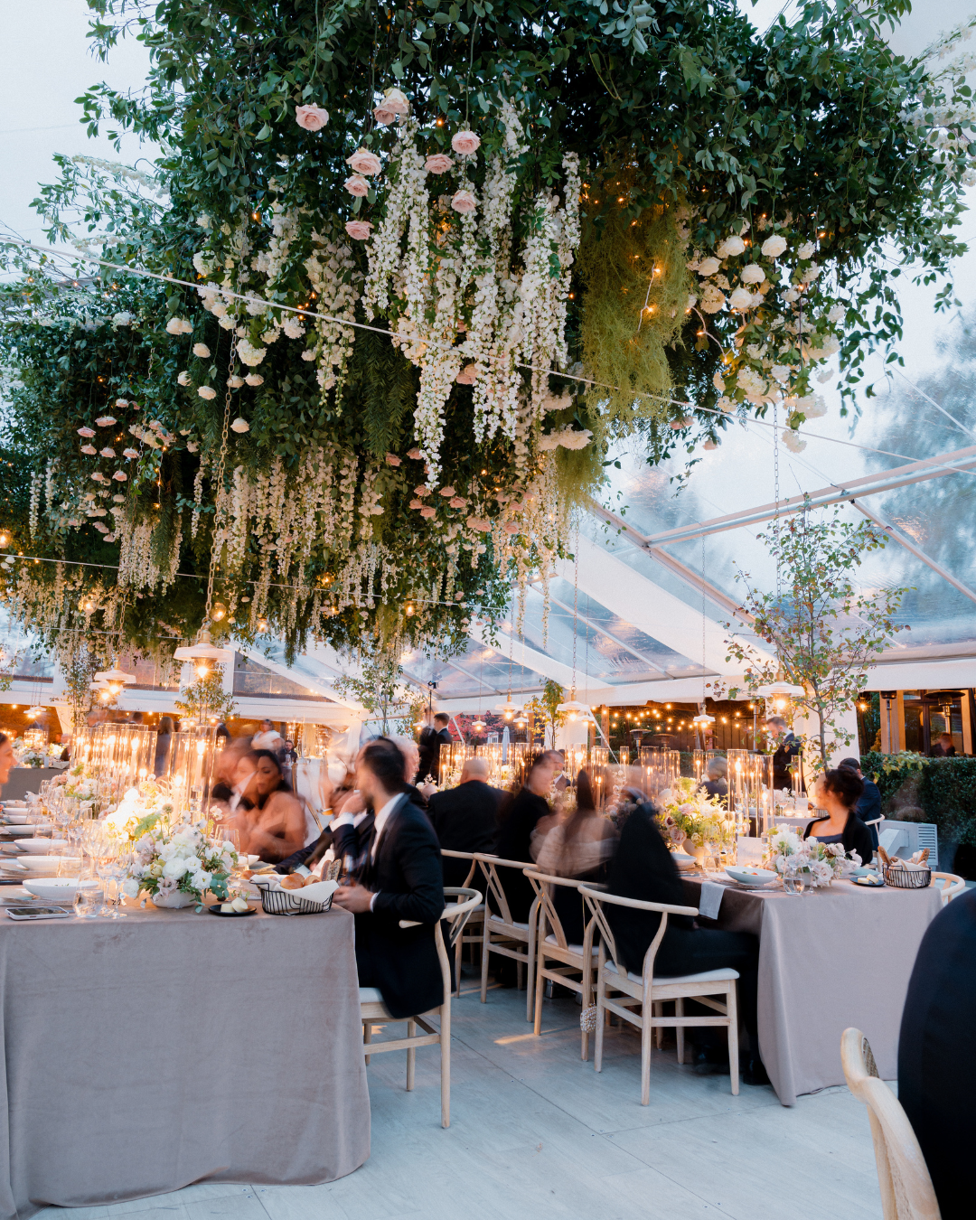 A tented reception with guests at tables underneath a hanging floral installation.
